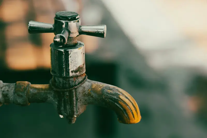 Close view of a weathered cross-handle outdoor tap with a stained spout and a drip forming underneath