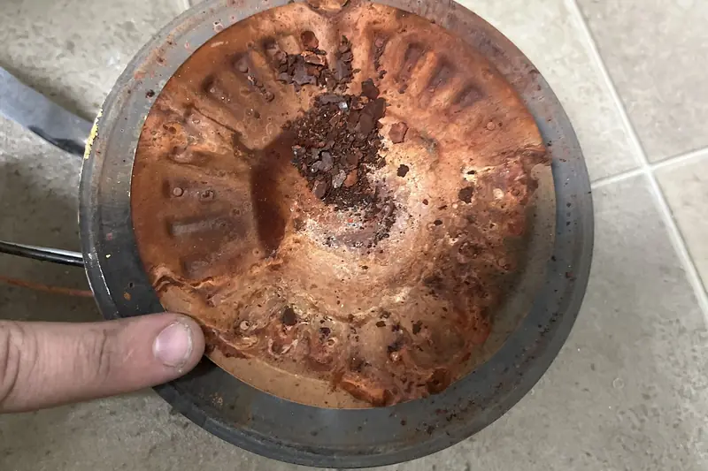 Close view of a corroded water heater base plate with a crust of reddish sediment, held over a tiled floor