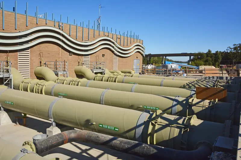 Rows of large pale green intake pipes and valves running alongside a brick pumping building under a clear blue sky